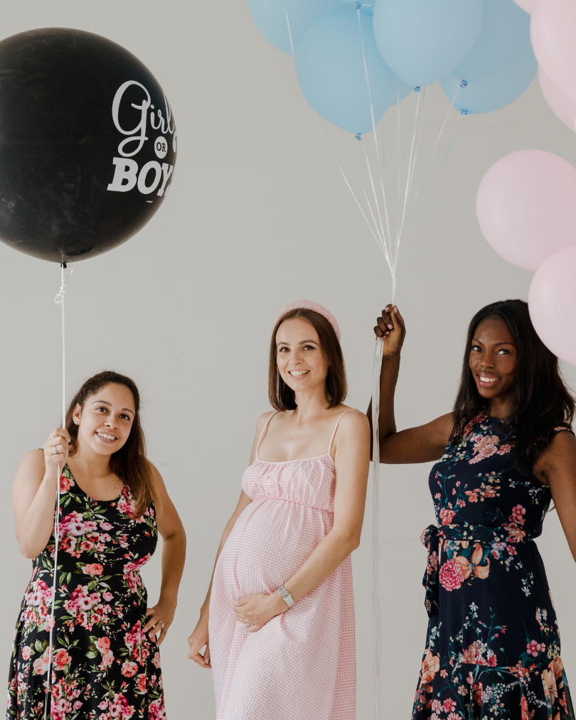 3 ladies standing for a photo pose with the middle woman being pregnant wearing a pink dress
